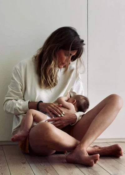 Frau in cremefarbenem Top hält Baby auf Holzboden im MILK AWAY Collection