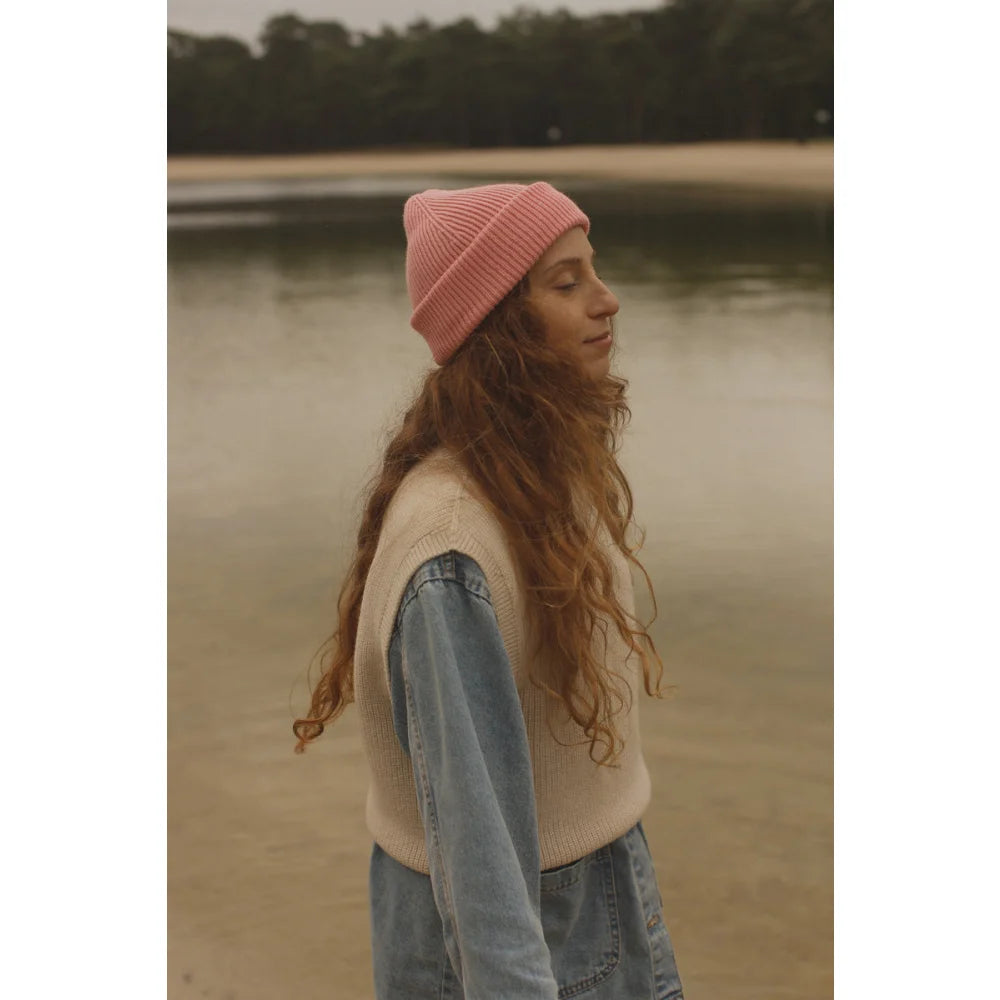 Junge Frau mit langen braunen Wellen, pinker Beanie, helles Sweatervest und Jeans