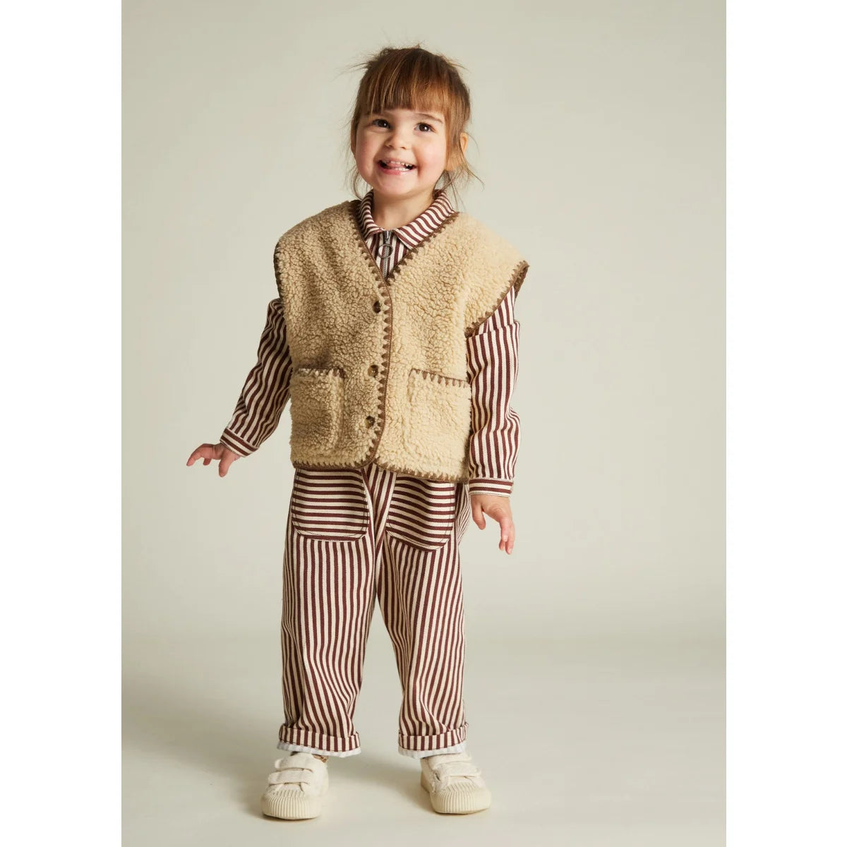Beige Shearlingweste Amos Folk Gilet Sand mit braunen Knöpfen und Taschen über gestreiftem Hemd