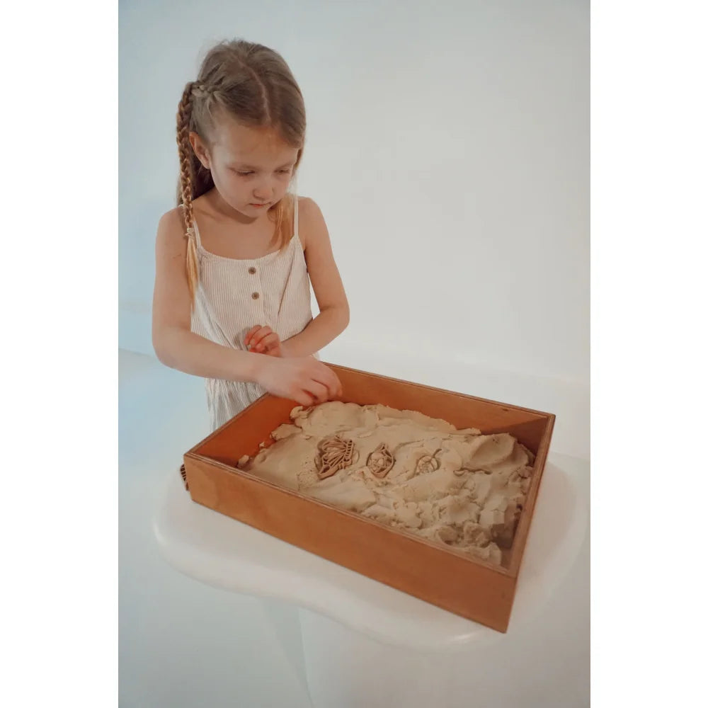 Junge Mädchen in hellem Kleid spielt mit Kinetic Sand im Holztablett für Ausstecher Schmetterlingszyklus