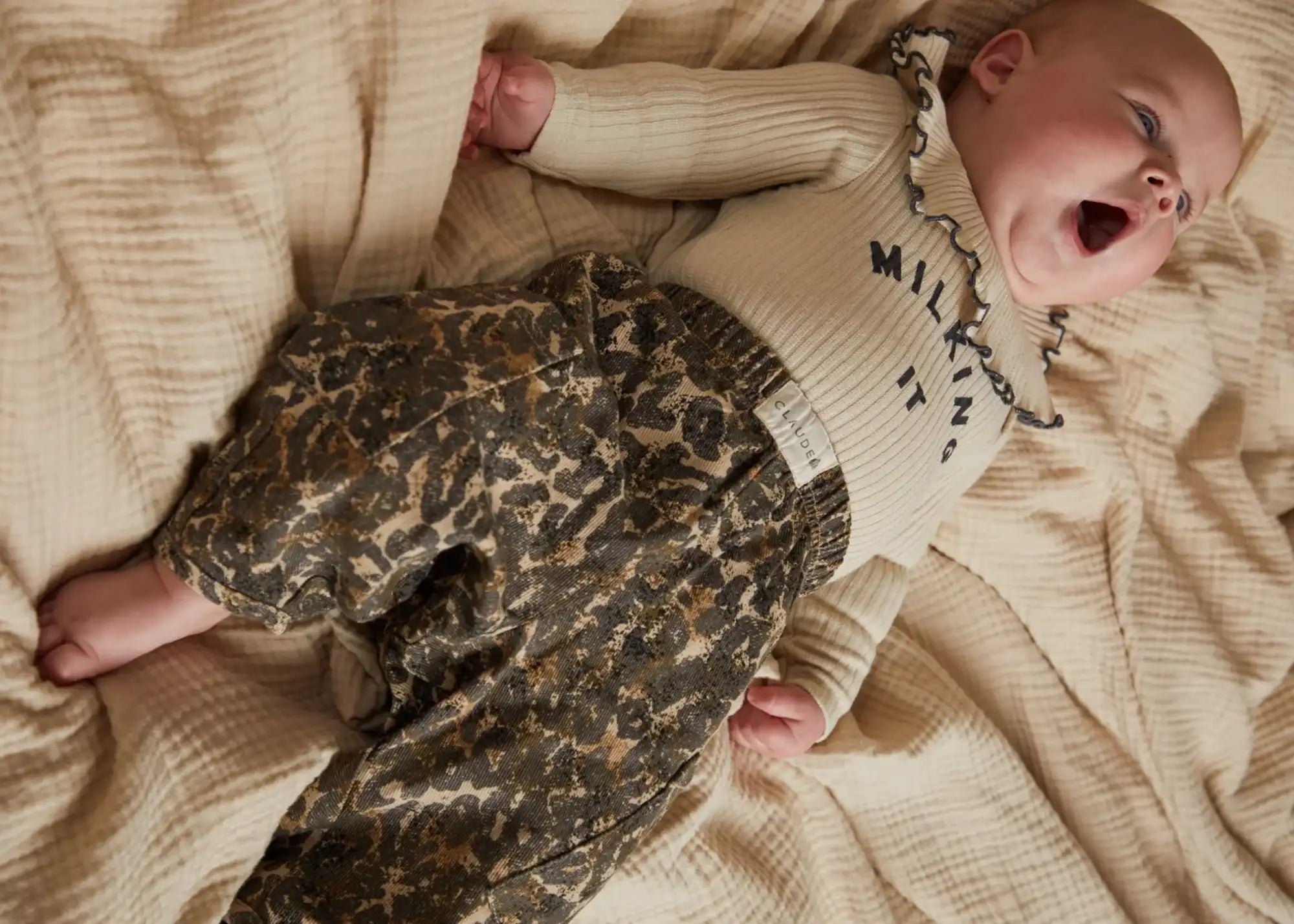 Das Baby trägt ein cremefarbenes, geripptes Langarmoberteil mit schwarzem besticktem Text und Rüschen am Ausschnitt, kombiniert mit Leopard-Print-Hosen in Schattierungen von Braun und Beige.