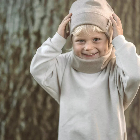 Lächelndes Kind mit beigem Hut und weißer Kleidung trägt Beanie rib strick sand