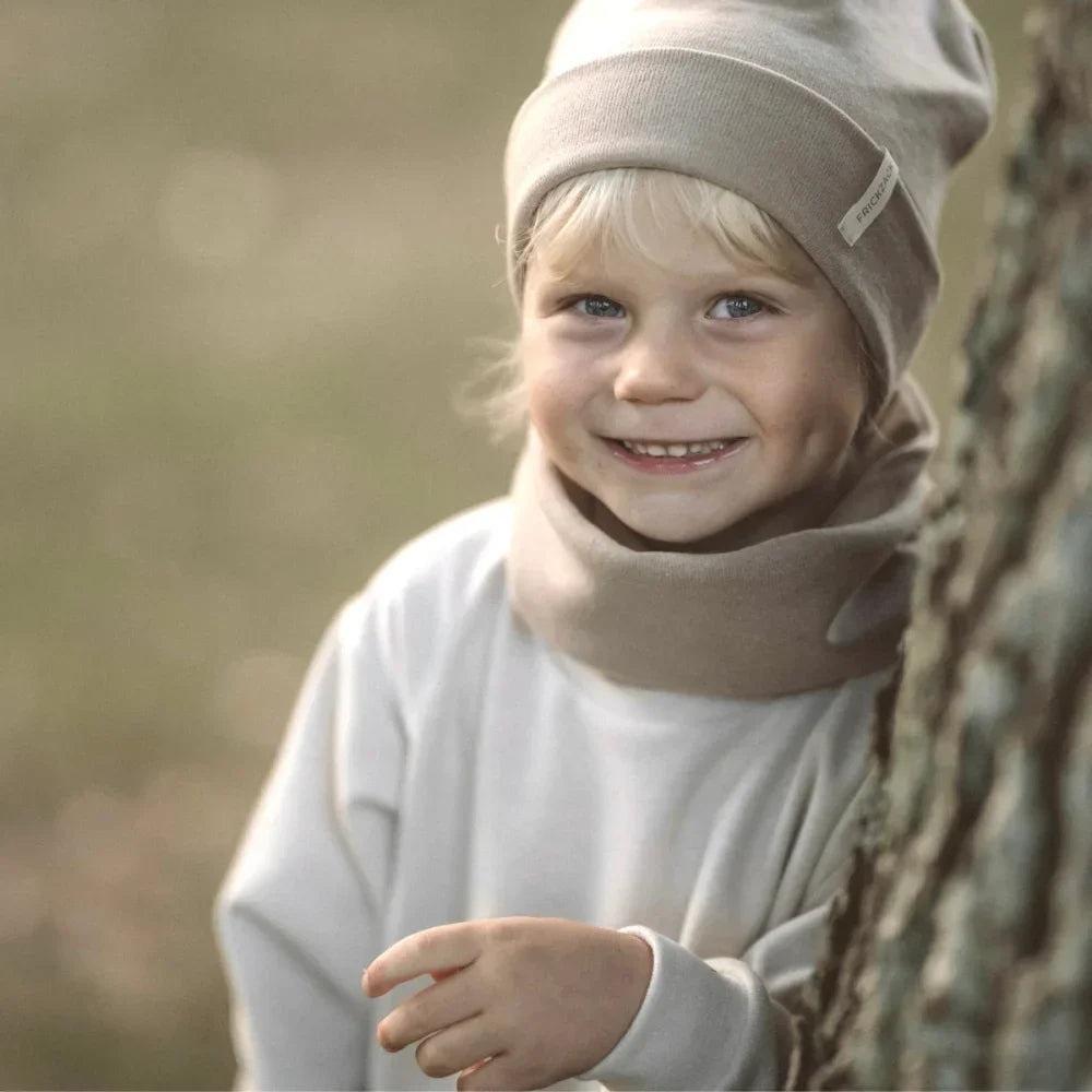 Lächelndes Kind mit weißem Hut und Pullover hinter einem Baum, Beanie Rib Strick sand