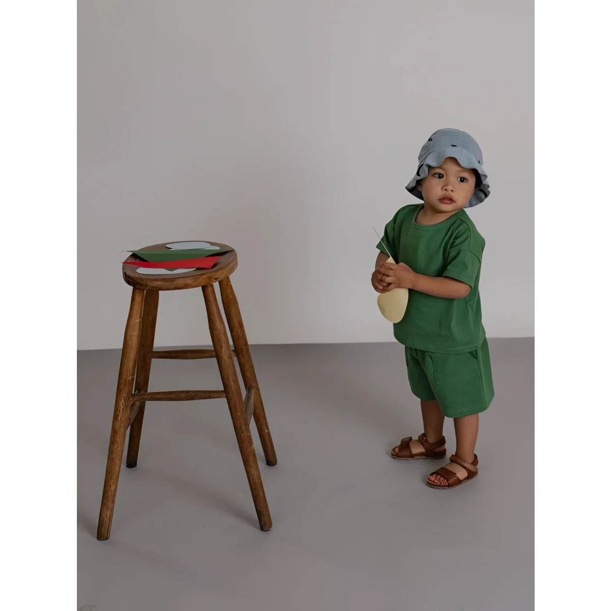 Ein kleinkind in einem grünen bermuda-outfit und hellblauem hut steht neben einem holzhocker mit bunten papierfiguren