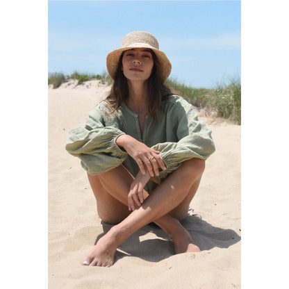 Frau in hellgrüner Tunika und Strohhut sitzt am Sandstrand, Produkt Bluse Lily Sage