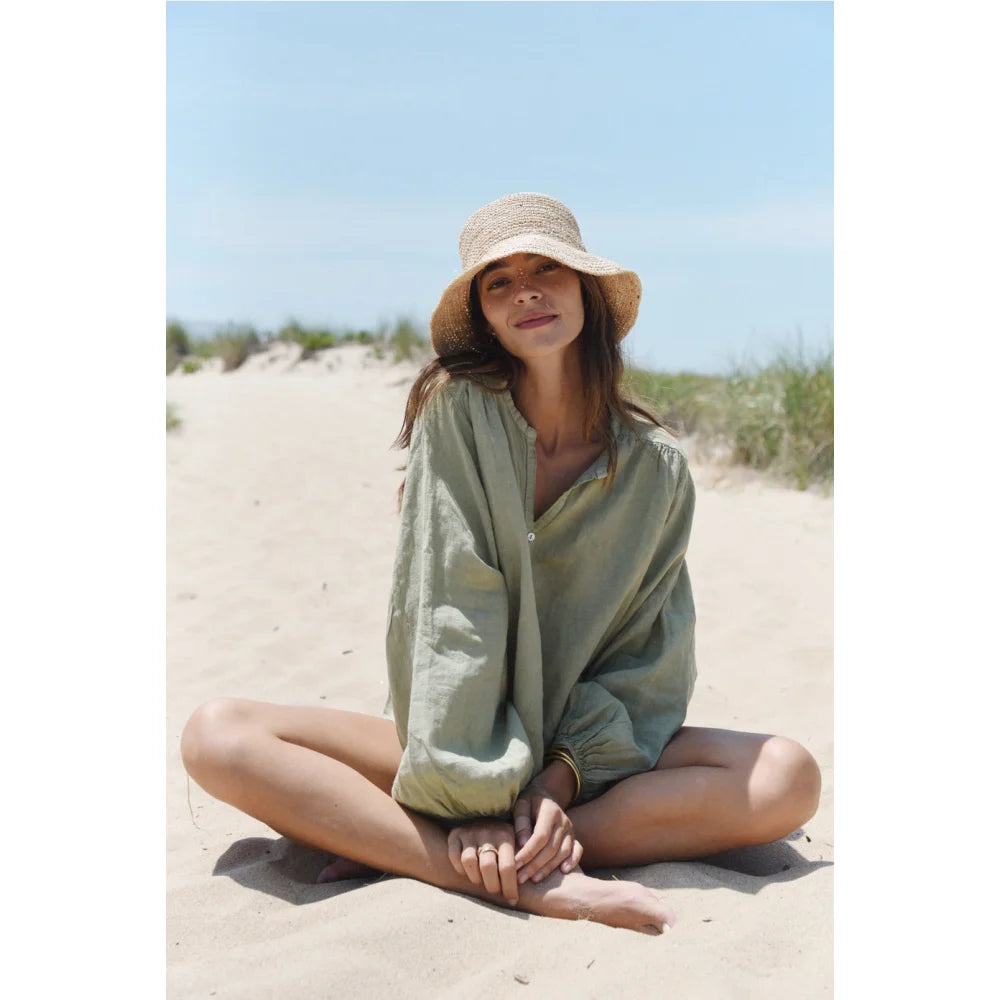 Frau in lockerem, hellgrünem Langarmshirt und Strohhut am Sandstrand sitzend