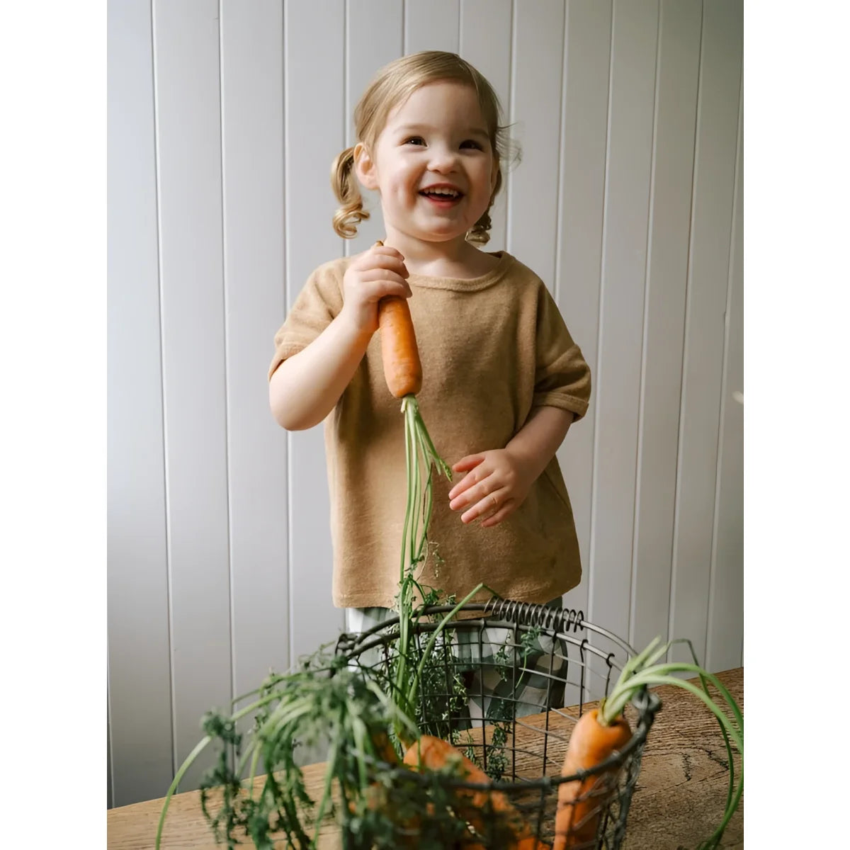Fröhliches Mädchen in Boxy T-Shirt Wheat Terry hält orange Karotte mit Grün