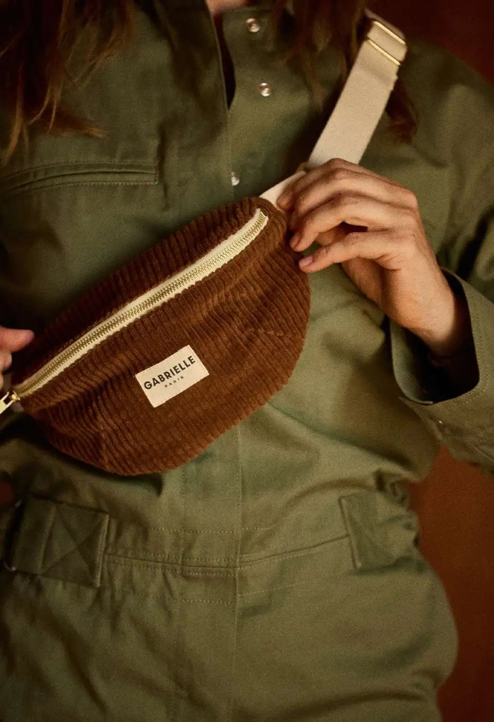 Braune Cord-Trendbauchtasche mit cremefarbenem Reißverschluss und einem weißen Label mit der Aufschrift „gabrielle paris“.