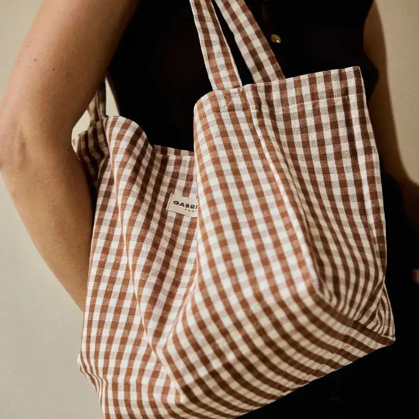 Eine braune und weiße Gingham-Karos Canvas-Tasche mit Henkel mit einem kleinen Markenlabel auf der Vorderseite.