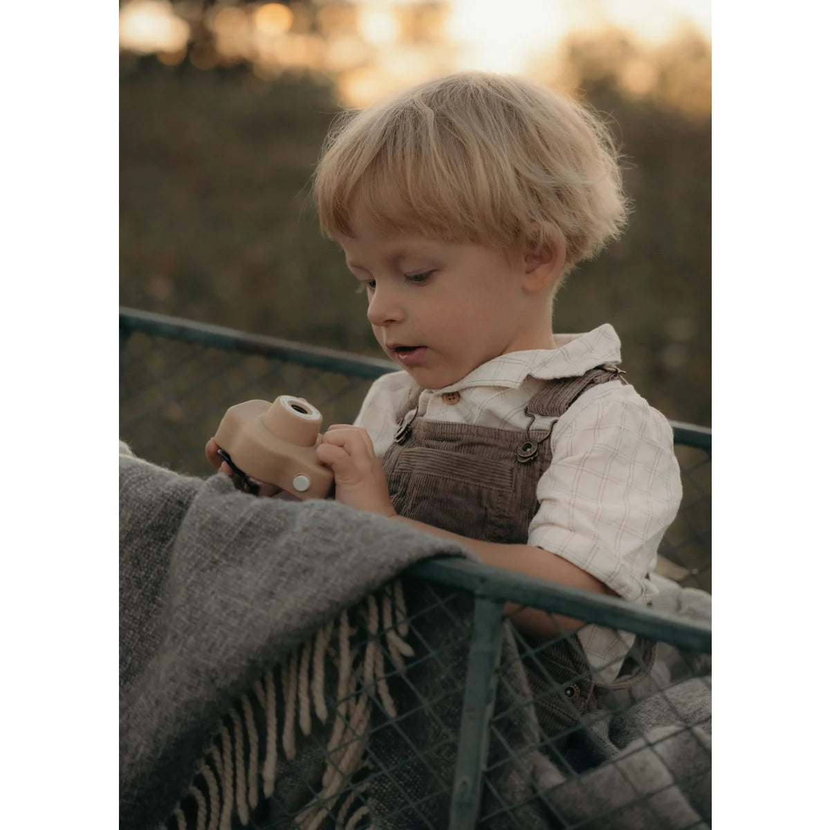 Ein kleines Kind in braunen Latzhosen hält eine Kamera auf einer Decke