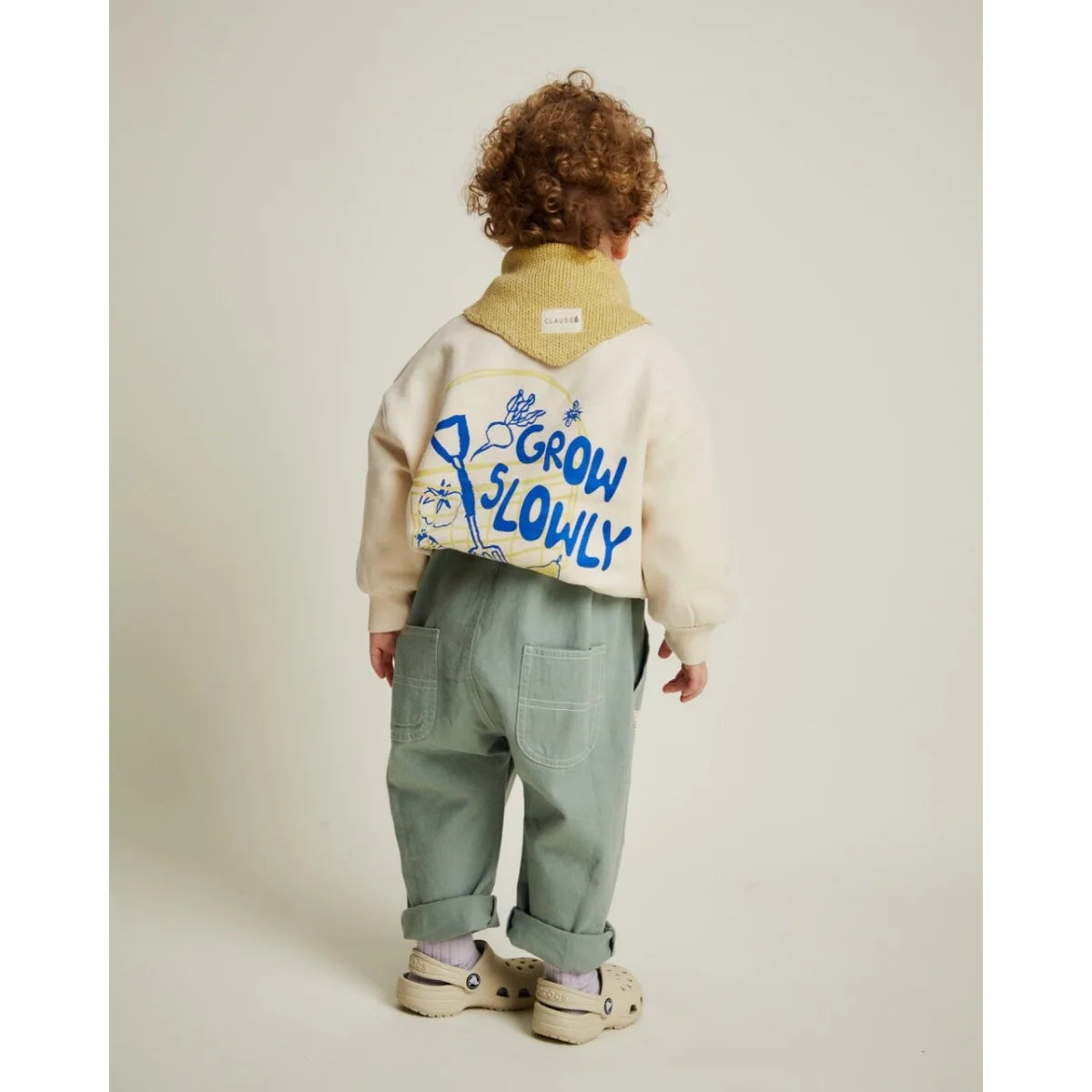 Kind trägt ein creamfarbener carner grow slowly sweater mit blauen buchstaben und hellgrüner cargohose