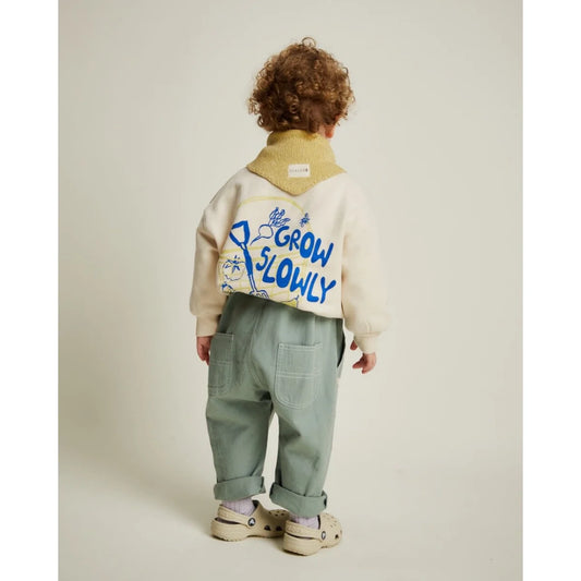 Kind trägt ein creamfarbener carner grow slowly sweater mit blauen buchstaben und hellgrüner cargohose