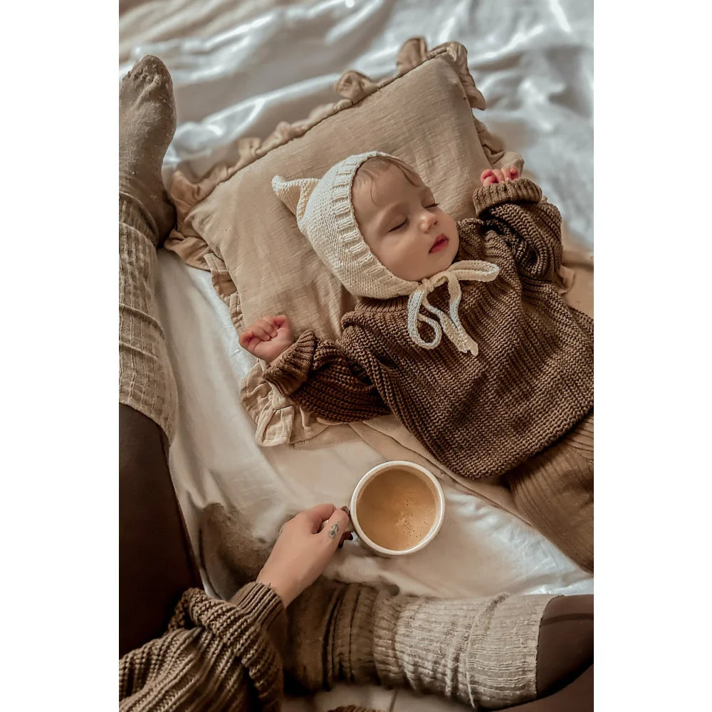 Baby in chunky knit coffee sweater and bonnet, resting on a soft pillow