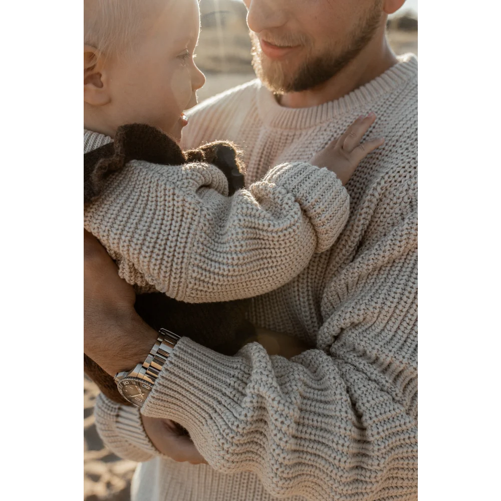 Erwachsener trägt einen Chunky Knit Sweater in Oat und hält ein Baby