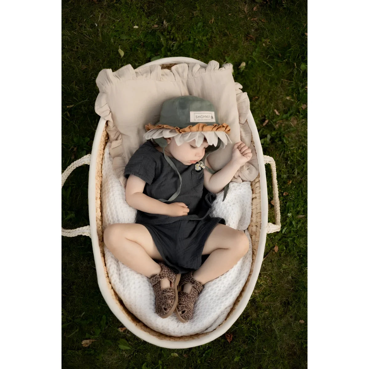 Ein baby trägt einen weichen grauen hut mit gerüschtem hellbraunem saum und sitzt in einem ovalen weidenkorb neben classic sandal cheetah sandalen