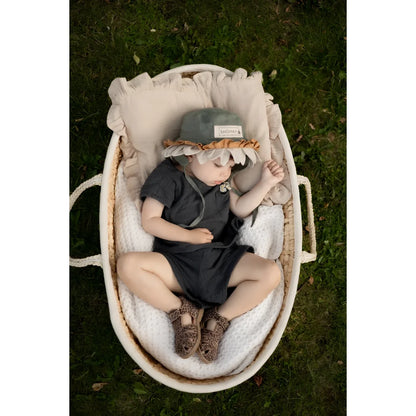Ein baby trägt einen weichen grauen hut mit gerüschtem hellbraunem saum und sitzt in einem ovalen weidenkorb neben classic sandal cheetah sandalen