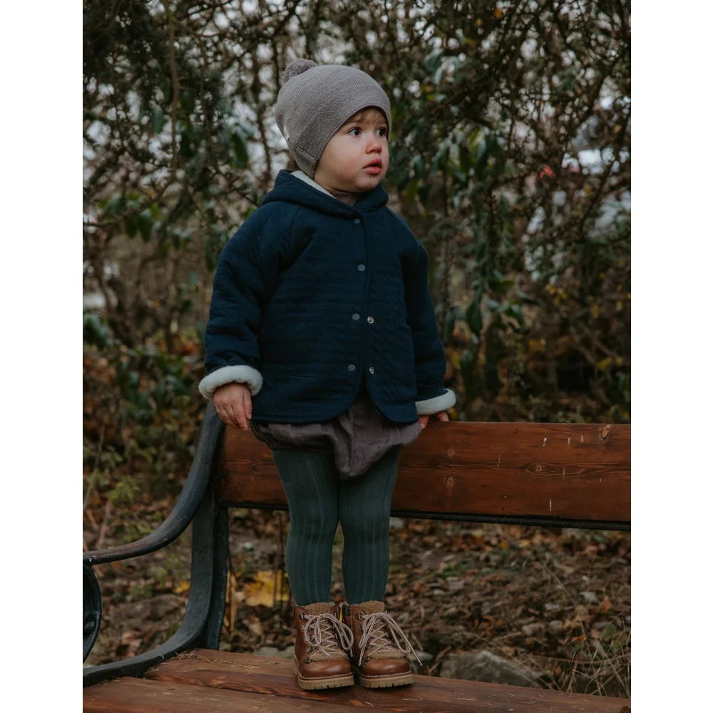 Kleiner Junge in dunkelblauer Winterjacke, grauem Mütze, grünen Strumpfhosen und braunen Stiefeln