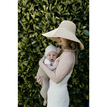 Frau mit Sonnenhut hält Baby vor blättrigem Hintergrund, präsentiert Cocoon Babydecke Sand