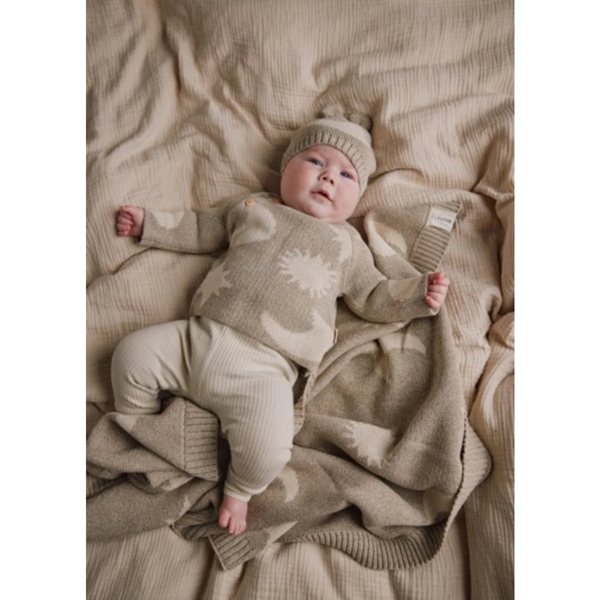 Baby in weichem Beige-Strickoutfit mit weißen Mond-Stern-Mustern auf passender Decke