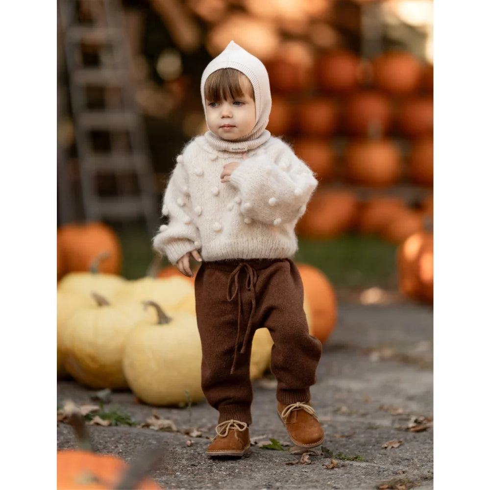 Kleiner Junge in cremefarbenem Pom-Pom-Pullover, braunen Hosen und passenden Schuhen