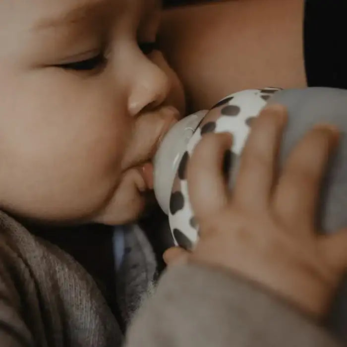 Die Hand eines Babys umfasst eine grau-weiße gepunktete Flasche mit einer weichen, abgerundeten Saugernippel.