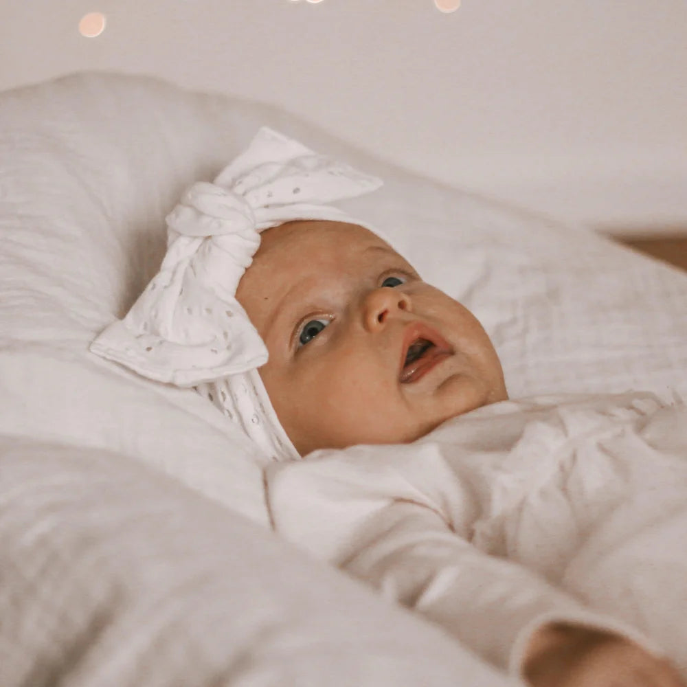 Baby in white outfit with bow headband on bedding showcasing Headband Milk Flowers