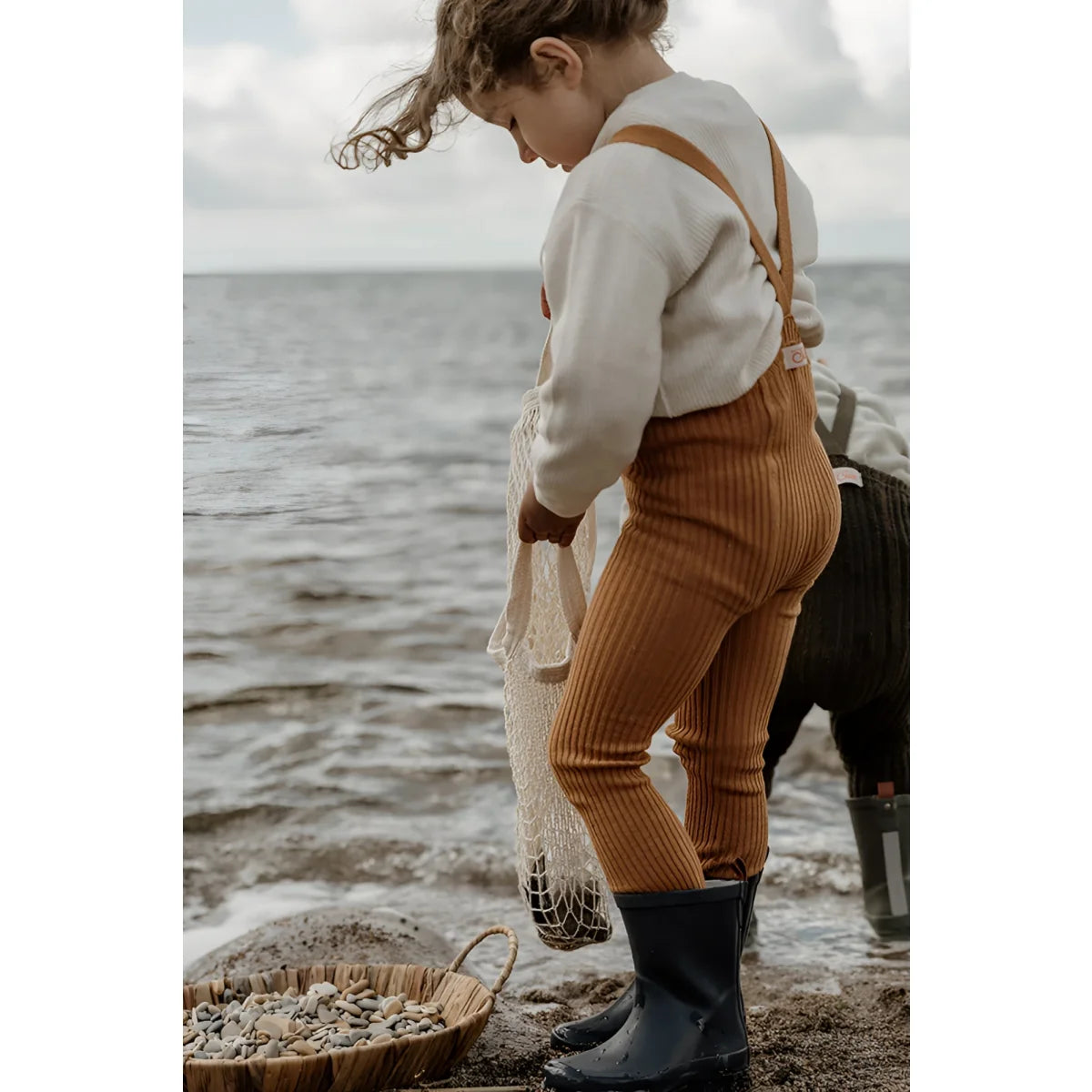 Kind mit cremigem Pullover, oranger Latzhose und dunklen Stiefeln am Wasser