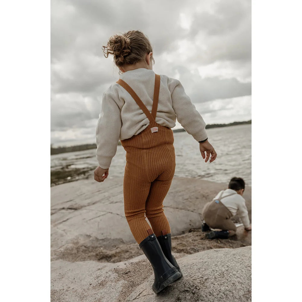 Kind in rostfarbenen Overalls und Gummistiefeln am Strand in Hippy Footless Strumpfhosen