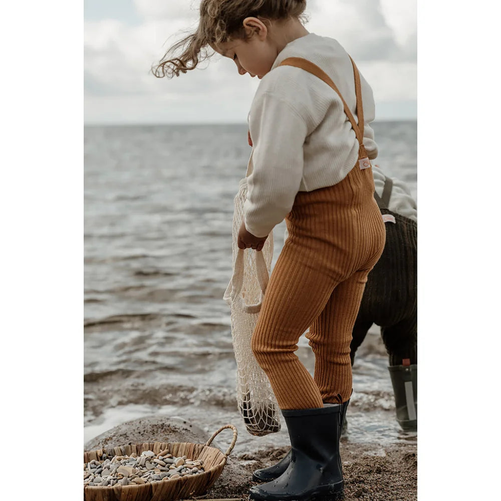 Kind in cremefarbenem Pullover, Cordhosen und Gummistiefeln am Strand