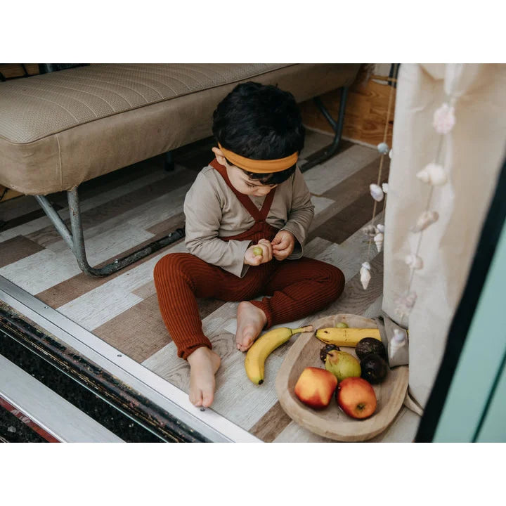 Kind in beigem Shirt, Zimt-Ripp-Hose mit Trägern und orange Headband