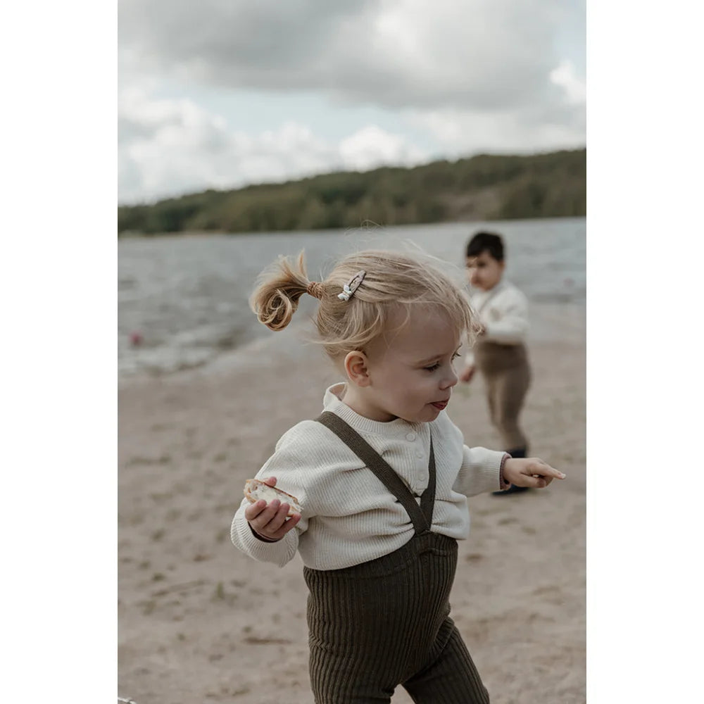 Kleinkind mit weißem Pullover und dunklen Latzhosen am Strand in Seaweed Brown