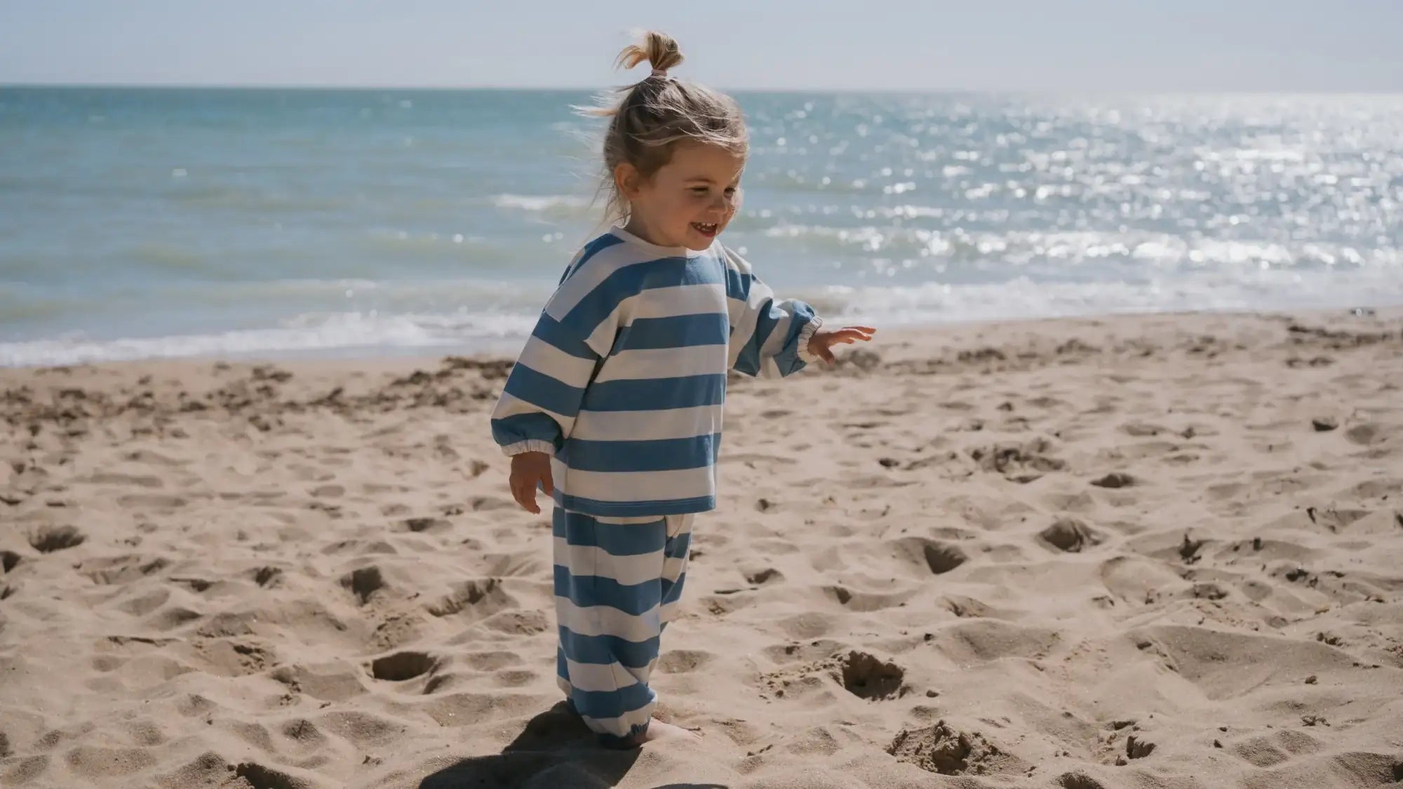 Ein junges Mädchen in einem blauen und weißen gestreiften, langärmligen Schlafanzug steht am sandigen Strand.