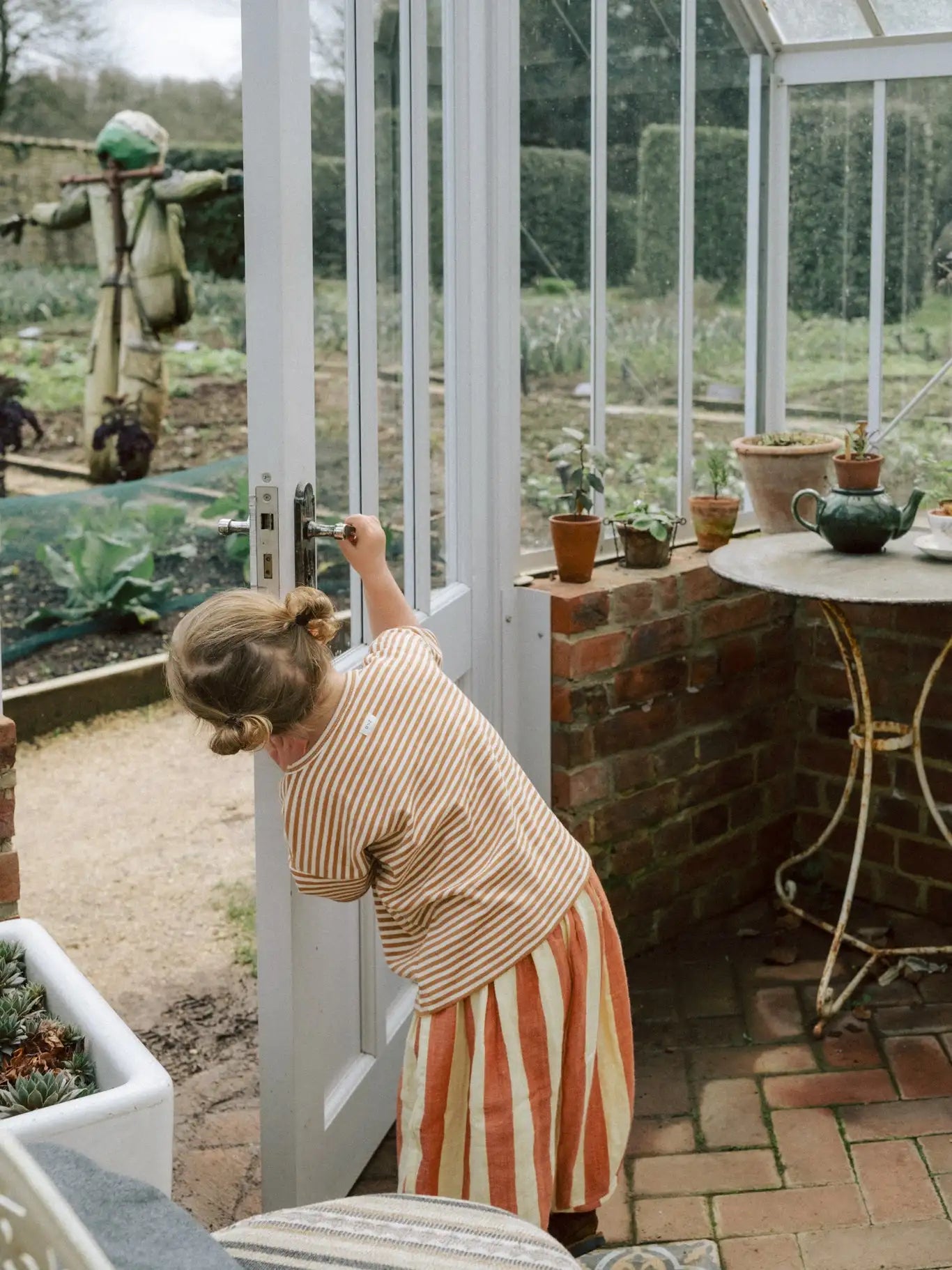 Ein junges Mädchen in einem gestreiften Oberteil und einem orange-weißen Rock steht an der Tür eines Gewächshauses, ihre Hand auf dem silbernen Türknauf.