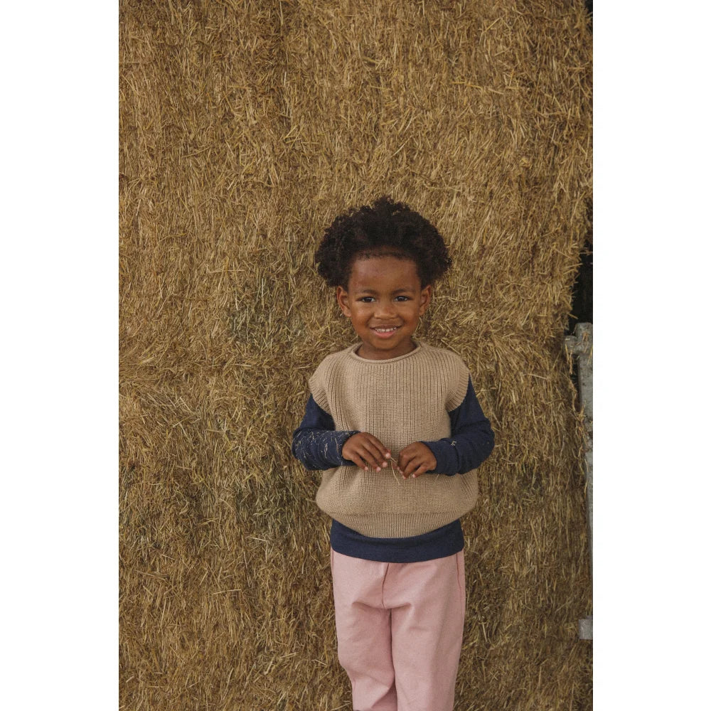 Junges Kind mit dunklen Locken trägt Sandfarbenes Kinderweste Harvey und pinke Hosen