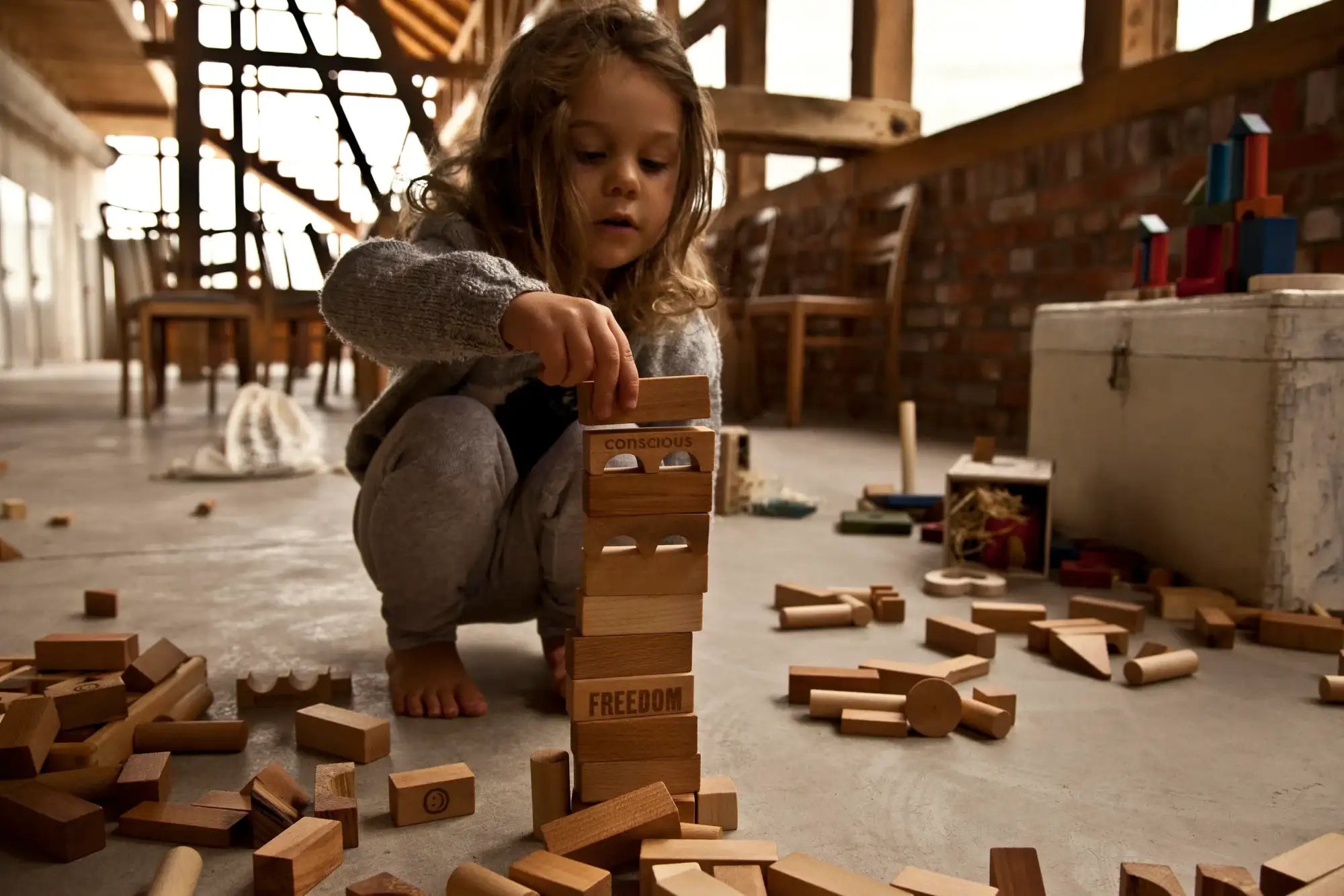 Ein Kind hockt sich hin und platziert vorsichtig einen Holzblock mit dem eingravierten Wort „Freiheit“ in einen hohen Turm aus ähnlichen Blöcken.