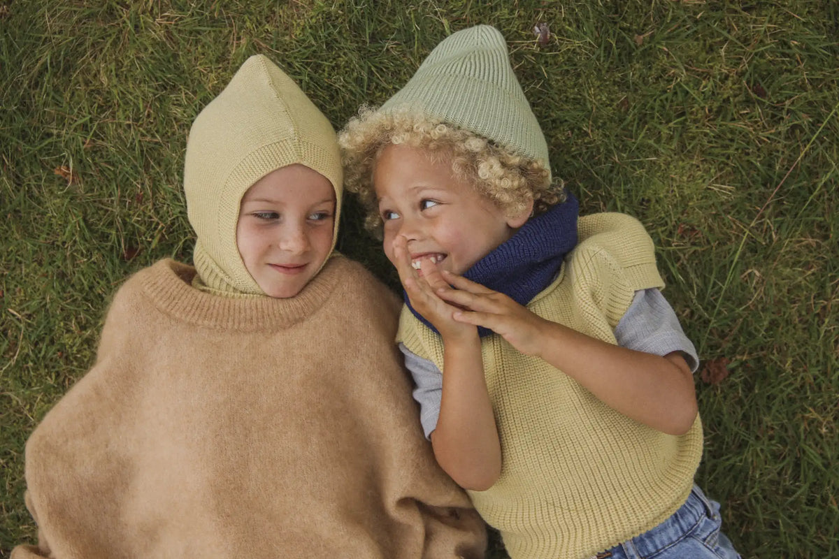 Zwei Kinder liegen auf dem Gras und tragen gestrickte Kapuzenmützen und gemütliche Strickkleidung.
