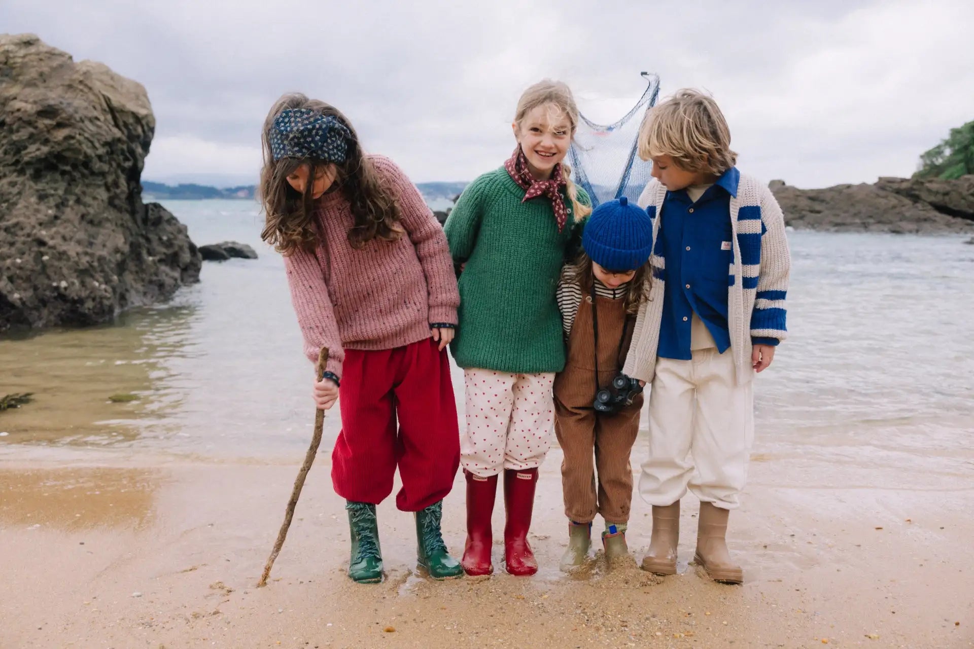 Vier Kinder stehen an einem sandigen Strand in farbenfroher, gemütlicher Strickkleidung und mit Regenstiefeln, nahe an Felsen und Wasser.