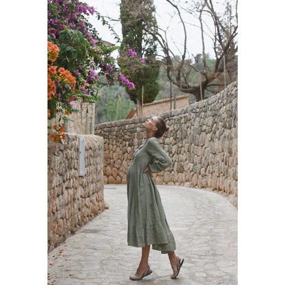 Eine frau trägt ein langes olivgrünes stufiges kleid mit lockeren ärmeln von kleid mara eucalyptus perfekt für ökologische baby- & kinderprodukte