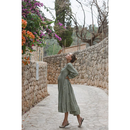 Eine frau trägt ein langes olivgrünes stufiges kleid mit lockeren ärmeln von kleid mara eucalyptus perfekt für ökologische baby- & kinderprodukte