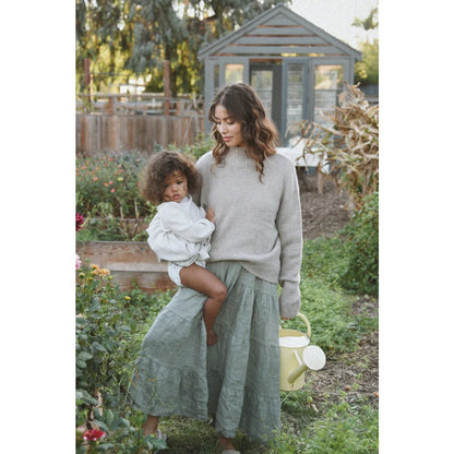 Eine frau trägt das nachhaltige kleid mara eucalyptus in salbeigrün mit einem kind und gießt mit einer gelben kanne nachhaltige babykleidung schweiz