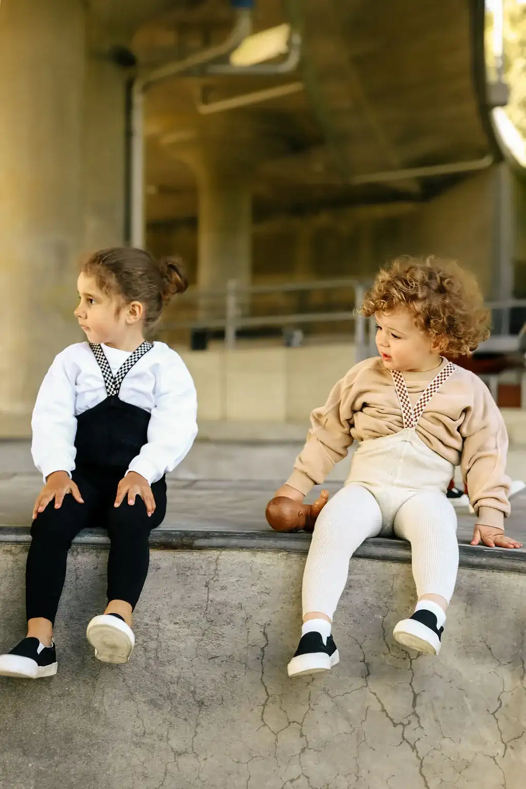 Zwei kleine Kinder sitzen nebeneinander auf einer Betonbrüstung, eines trägt schwarze Latzhosen mit weißen langen Ärmeln, das andere beigen Overalls, die mit einem passenden Sweatshirt kombiniert sind.