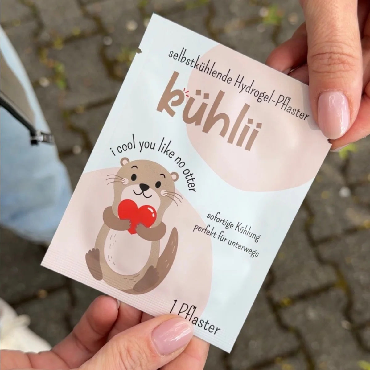 Ein süßer beige brauner cartoon-otter hält ein rotes herz auf einer weißen selbstkühlenden hydrogelpflaster-packung von kühlІІ