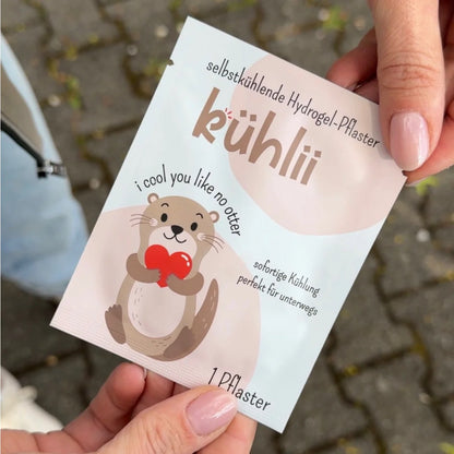 Ein süßer beige brauner cartoon-otter hält ein rotes herz auf einer weißen selbstkühlenden hydrogelpflaster-packung von kühlІІ