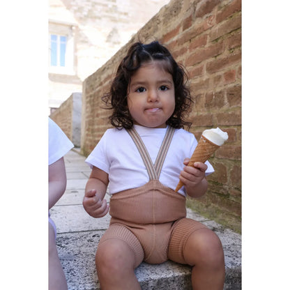 Ein kind trägt kurze strumpfhose mit träger in hellbraun gestreift und beige gestrickte overalls hält eine vanilleeistüte nachhaltige babykleidung schweiz