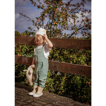 Kind mit hellgrünen Latzhosen, weißer Strickmütze und beigefarbenen Gummistiefeln hält kleines Kuscheltier Elefant aus Leinen