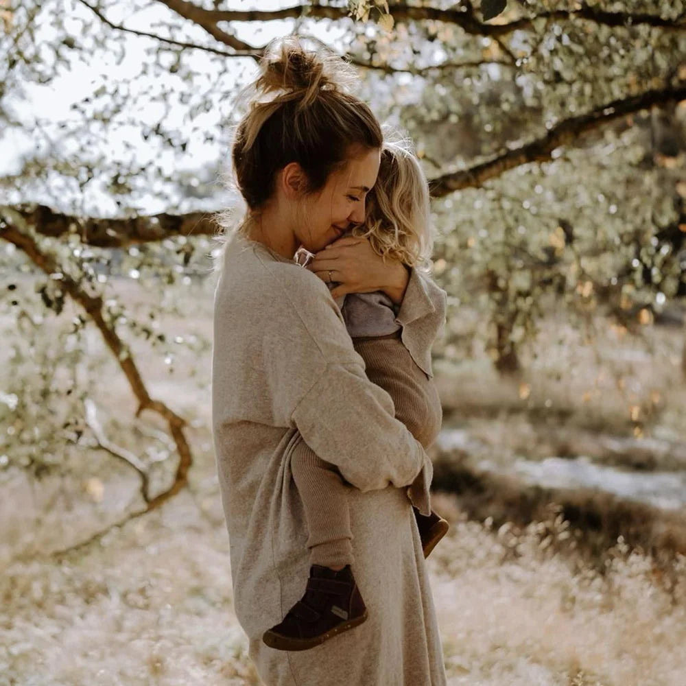 Frau in beigem Kuscheltier-Schneeeule-Pulli umarmt Kind im Schatten der Bäume