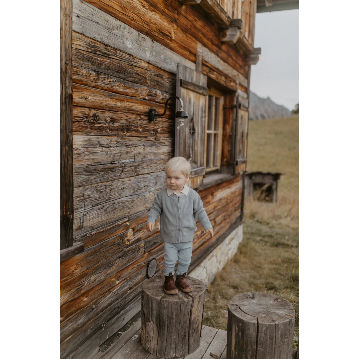 Kleines Kind in nachhaltiger Babykleidung Schweiz auf Holzstumpf vor Hütte