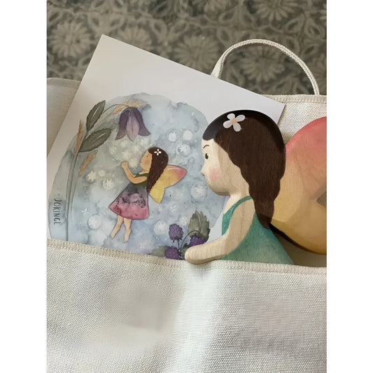 Eine holzfee mit braunen haaren grünem kleid und pfirsichfarbenen flügeln in einer beige tasche aus nachhaltiger babykleidung schweiz