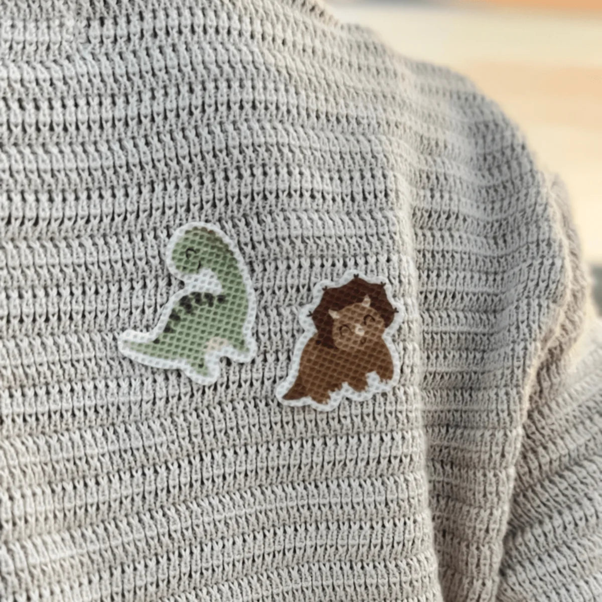Zwei gestickte Dino-Patches, grün und braun mit weißer Umrandung, auf grauem Strickpullover der nachhaltigen Babykleidung Schweiz Mückenschutz DINO Edition