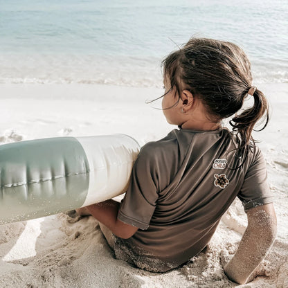 Ein kind trägt einen braunen kurzärmligen rash guard mit ocean child logo und turtle patch am strand mit grün-weißem luftreifen